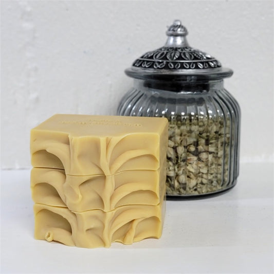A stack of three golden rectangular bars of soap with textured tops, displayed next to a ribbed clear glass jar filled with dried botanicals on a white surface.