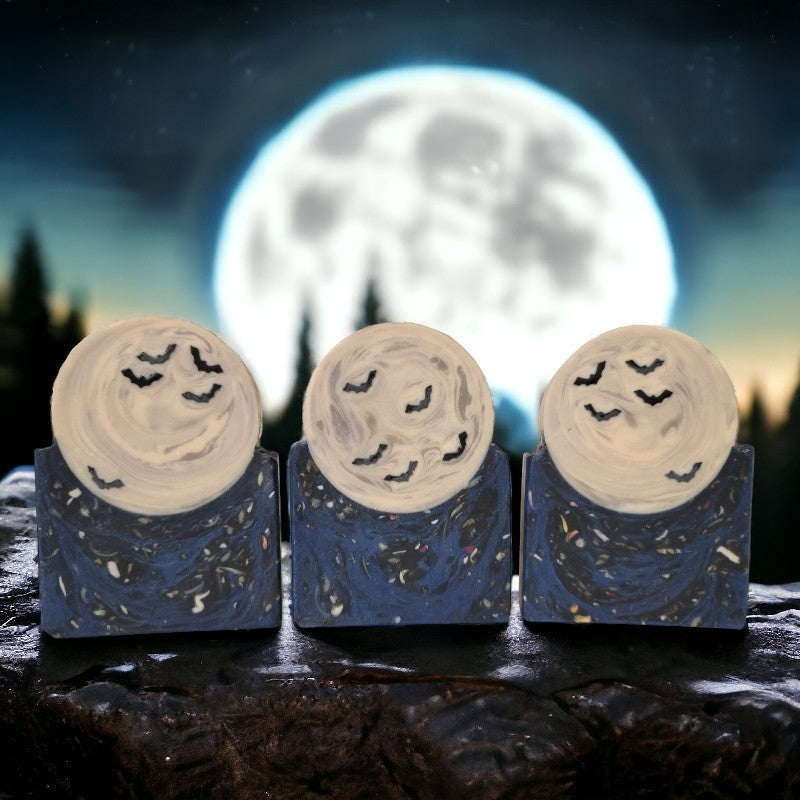 Dark blue speckled soap base with a full moon and bats sitting on it. These soaps are sitting on a stone surface in front of a rising full moon.