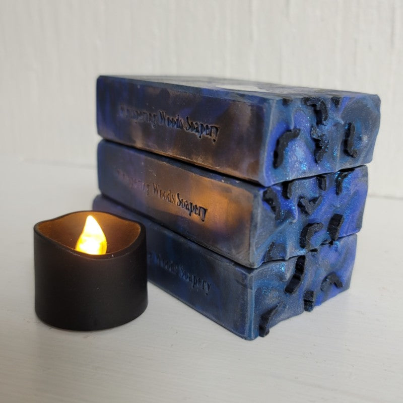 Three blue soap bars, stamped 'Whispering Woods Soapery,' stacked on a white surface by a black tealight candle. The tops of the soap have blue sparkle and small black soap-dough bats.