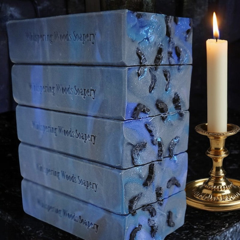 A stack of blue bars of soap with small bats and blue glitter sit atop a black counter with a lit candlestick beside them