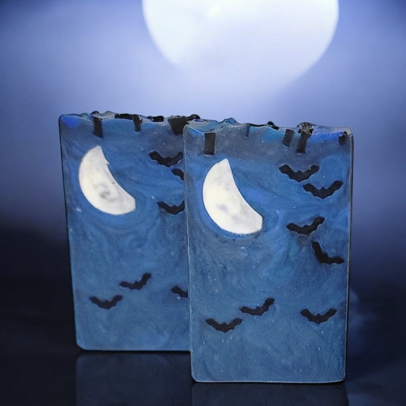 Two blue bars of soap with seven small bats and a crescent moon embed on a black glass surface with bluish fog in the background
