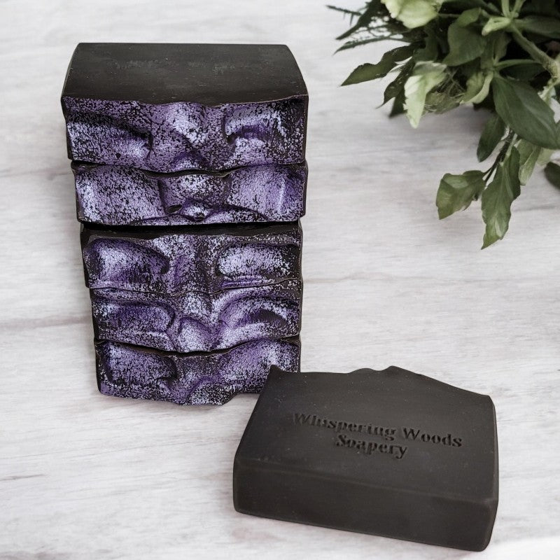 Six bars of black soap with light purple tops are stacked on a marble counter