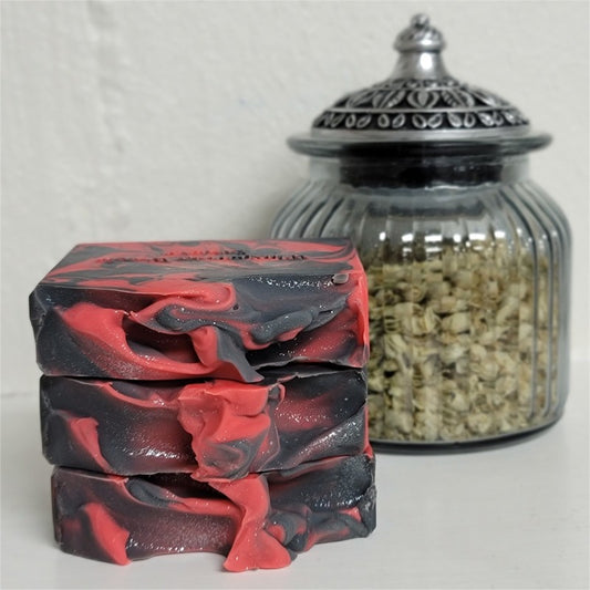 Three stacked rectangular bars of soap with a black and red marbled pattern, displayed next to a ribbed glass jar filled with dried botanicals on a white surface.