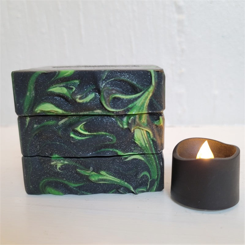 Three stacked rectangular bars of soap with a black and green marbled swirl pattern, displayed next to a small black candle on a white surface against a white textured wall.