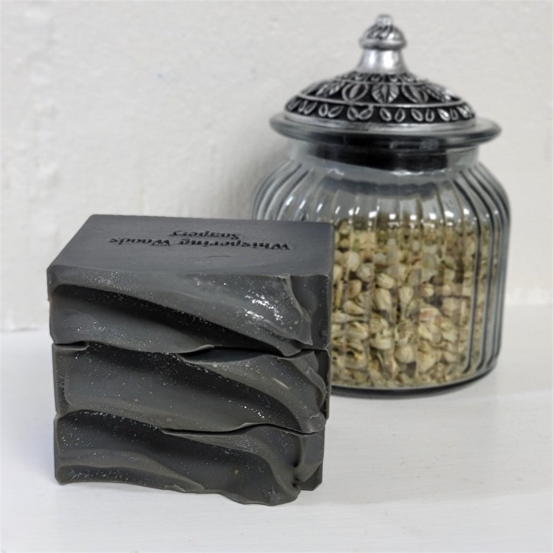 A stack of light black soap bars with 'Whispering Woods Soapery' stamped on the top bar, displayed next to a glass jar filled with dried botanicals on a white surface.