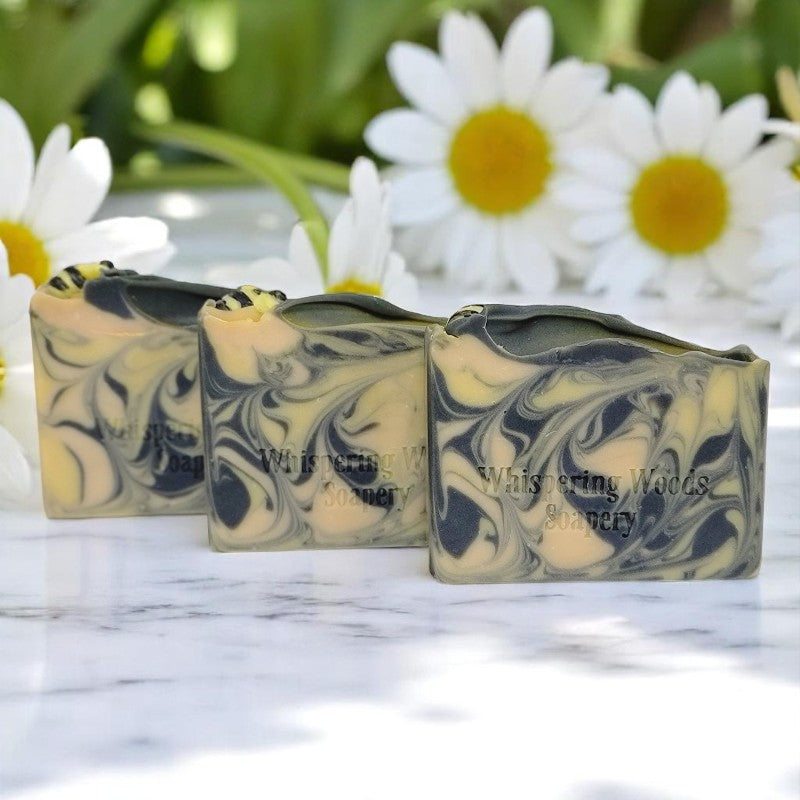 Three bars of soap with yellow, gold and black swirls that have tiny soap bees on top, on a marble bench with daisies in the background