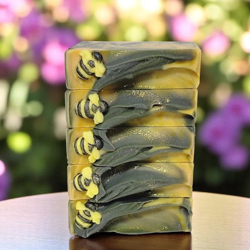 Five bars of soap with yellow, gold and black swirls that have tiny soap bees on top are stacked on a wood table with sunlit flower blurred behind them