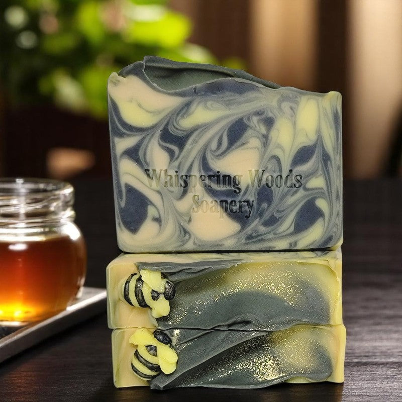 Three bars of soap with yellow, gold and black swirls that have tiny soap bees on top sit on a dark table with a jar of honey