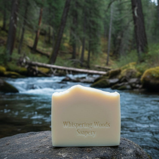 A bar of cream-colored soap with 'Whispering Woods Soapery' branding sits on a rock by a stream running through a woods
