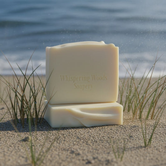 Two bars of cream-colored soap from 'Whispering Woods Soapery' on a sandy beach with grass