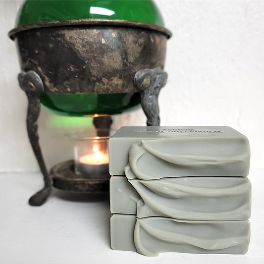 Three stacked rectangular bars of light gray soap with a textured, ridged top surface, displayed next to a vintage warmer with a lit tealight candle on a white surface against a white wall.