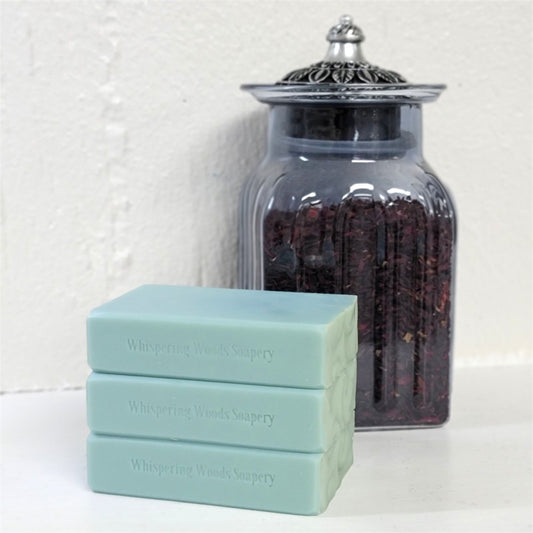 Three stacked duck-egg-blue soap bars labeled 'Whispering Woods Soapery' in front of a clear glass jar filled with dried hibiscus petals on a white surface against a white wall.