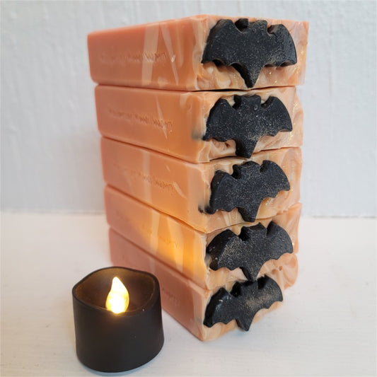 Five orange and cream bars of soap, stamped 'Whispering Woods Soapery,' stacked on a white surface near a black tealight. There are black soap bats on top of the bars.
