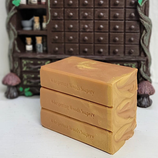 Three stacked rectangular bars of soap in a warm orange color, stamped with the brand name 'Whispering Woods Soapery,' set against a blurred dark brown wooden cabinet background.