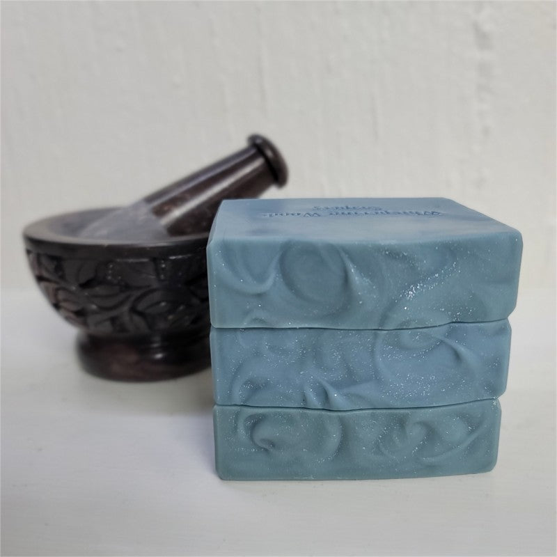 A stack of dusky-blue soap with sparkly textured tops next to a soapstone mortar and pestle on a white background.