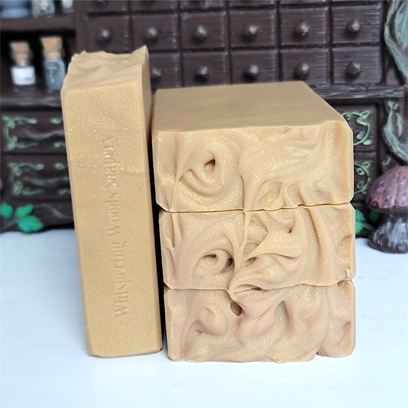Four bars of handmade soap in a warm honey color, with one bar standing vertically showing the stamped brand name 'Whispering Woods Soapery.' The other three bars are stacked to show the textured tops.