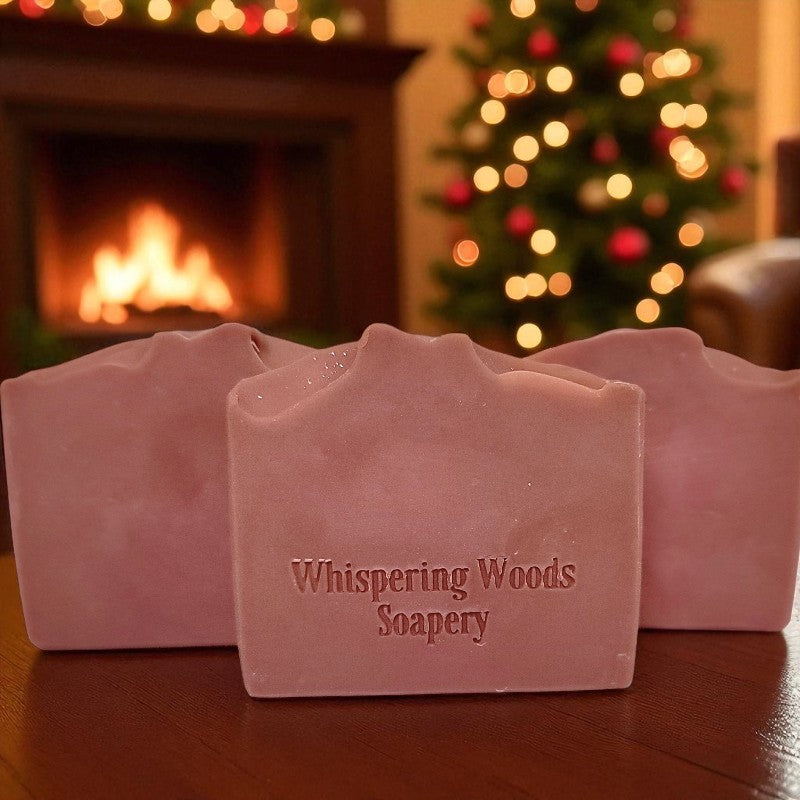 Three bars of soap with 'Whispering Woods Soapery' branding in front of a fireplace and Christmas tree