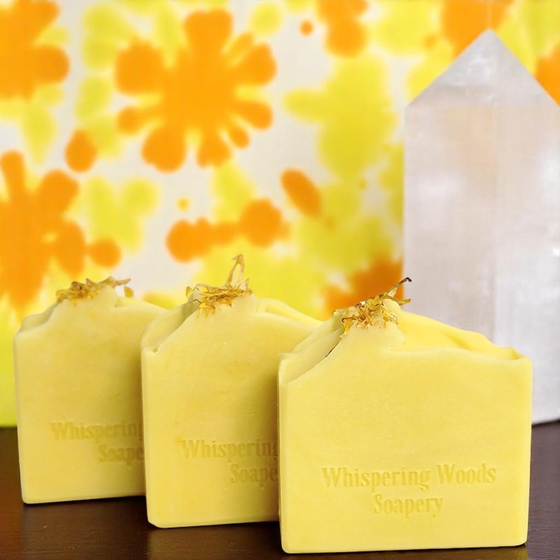 Three bars of yellow soap with dried calendula on top, on a dark table with orange and yellow tie-dye cloth in the background