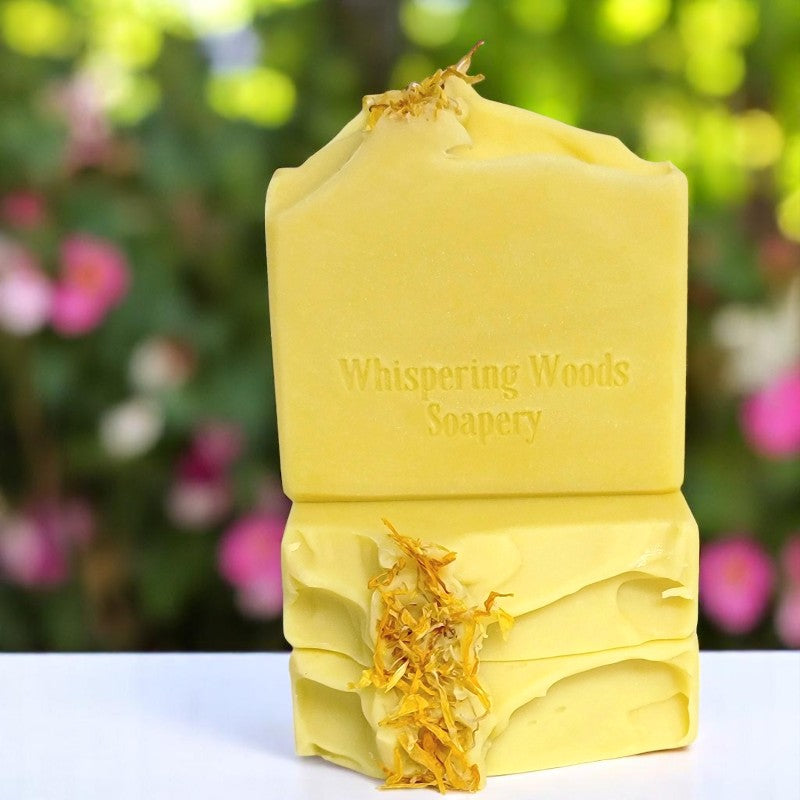 Three bars of yellow soap with dried calendula on top on a white bench with flowers in the background