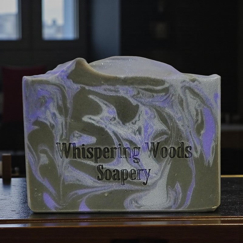 Silver, purple and black swirled soap bar with 'Whispering Woods Soapery' branding on a dark surface