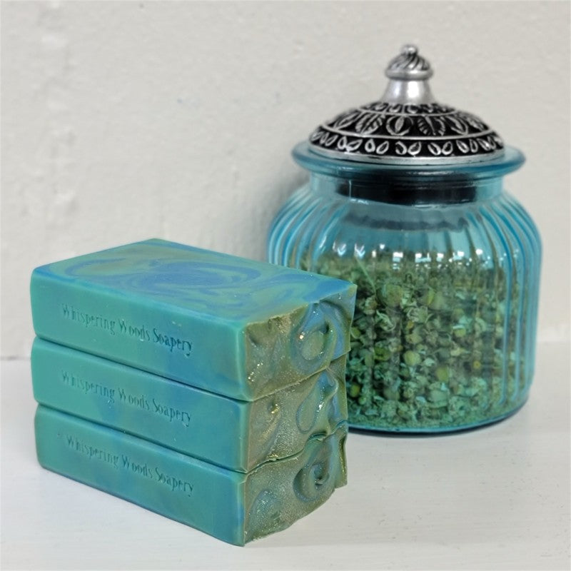 Three stacked turquoise and blue soap bars with swirled tops and gold sparkle, stamped 'Whispering Woods Soapery,' displayed next to a ribbed blue glass jar filled with dried herbs on a white surface.