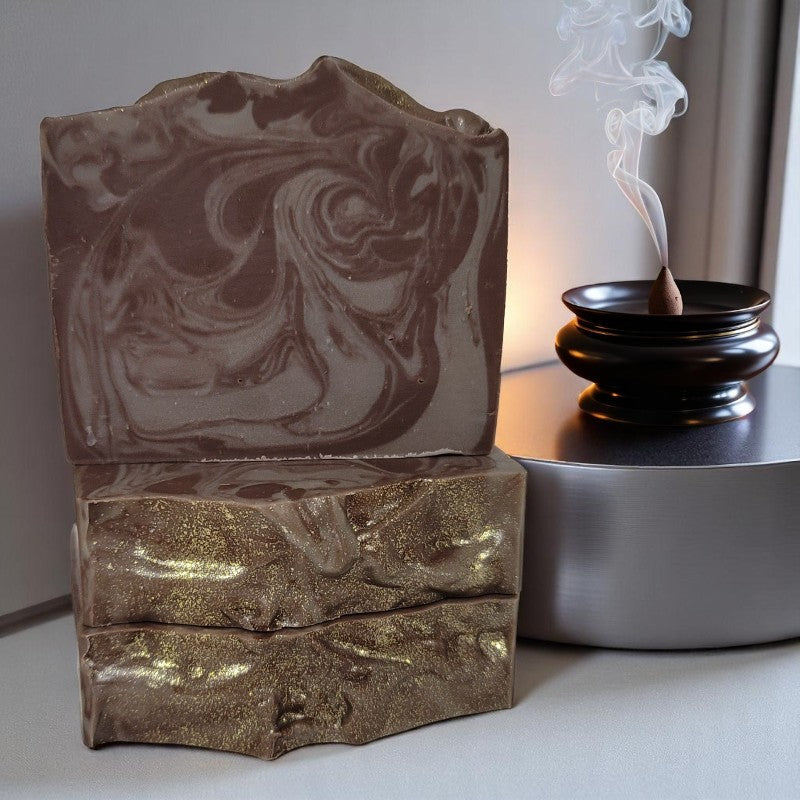Three bars of brown-swirled soap with sparkly gold tops on a counter with a cone of incense burning beside them