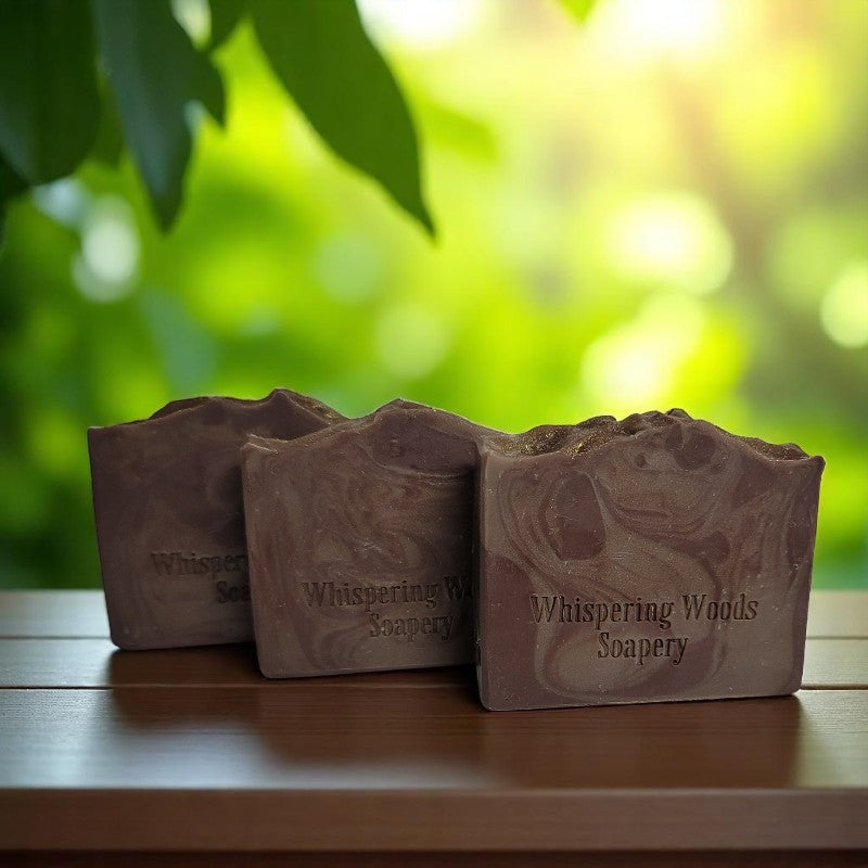 Three bars of brown-swirled soap with sparkly gold tops on a bench with sunshine filtering through green plants in the background