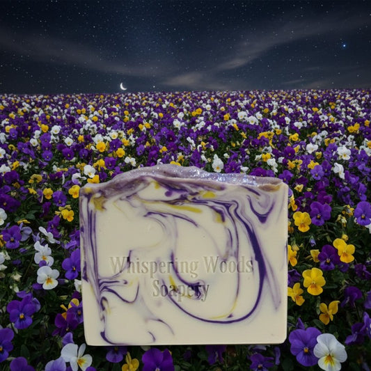 Bar of soap with 'Whispering Woods Soapery' branding in a field of purple and yellow flowers under a starry night sky
