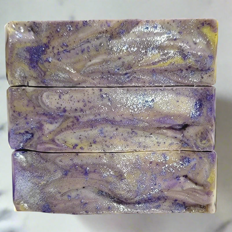 Close-up of three bars of purple white and yellow soap with sparkle on top sitting on a white background