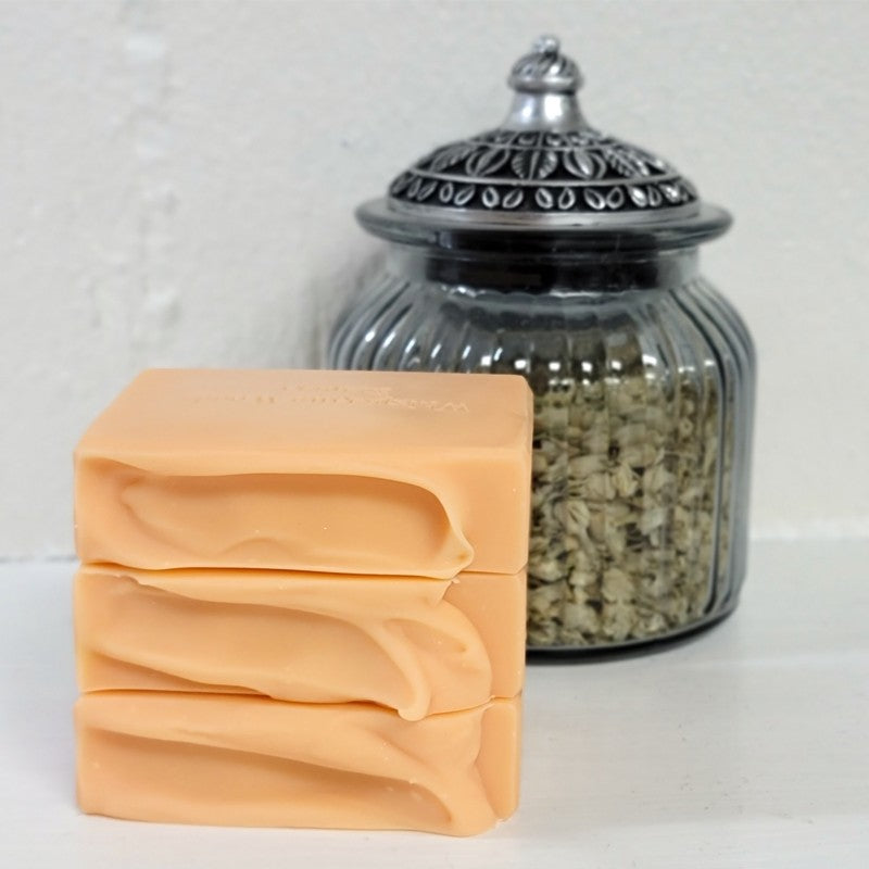Three stacked rectangular bars of pale peach-colored soap in front of a ribbed clear glass jar with a silver-toned lid, set against a white textured background.