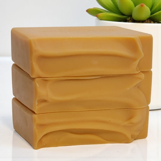 Three bars of orangish soap stacked on a white surface with a green plant in the background