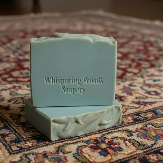 Two blue-green bars of soap with 'Whispering Woods Soapery' branding sit upon a persian silk rug