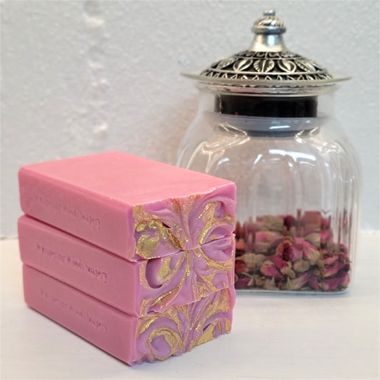 Three pink handmade soap bars from Whispering Woods Soapery stacked on top of each other, featuring a decorative gold mica swirl on their tops. A clear glass jar with a silver lid containing dried rose petals sits in the background against a white textured wall.