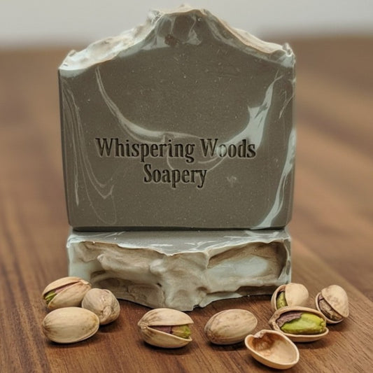 Two bars of soap with the branding 'Whispering Woods Soapery' sit on a wood surface with some pistachios scattered about