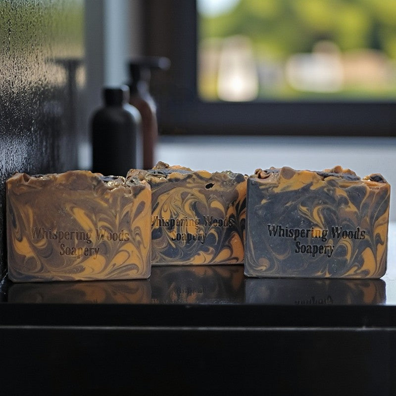 Three bars of soap with marbled design and 'Whispering Woods Soapery' branding on a reflective surface