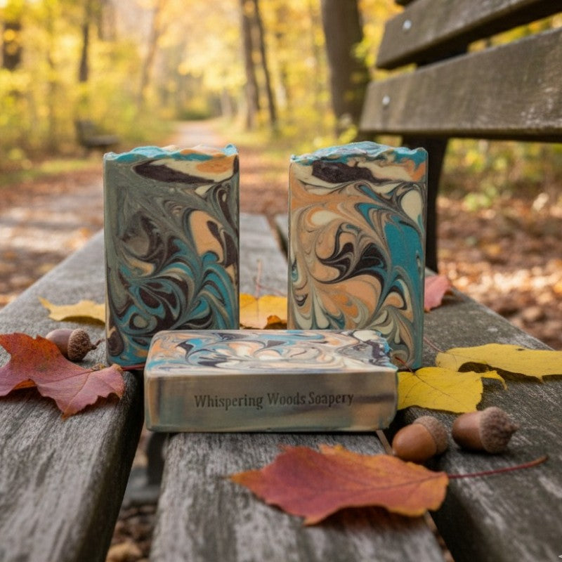Three marbled soap bars with 'Whispering Woods Soapery' branding on a wooden bench with autumn leaves and trees in the background
