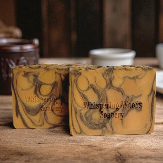 Two bars of soap with marbled design and 'Whispering Woods Soapery' branding on a wooden surface
