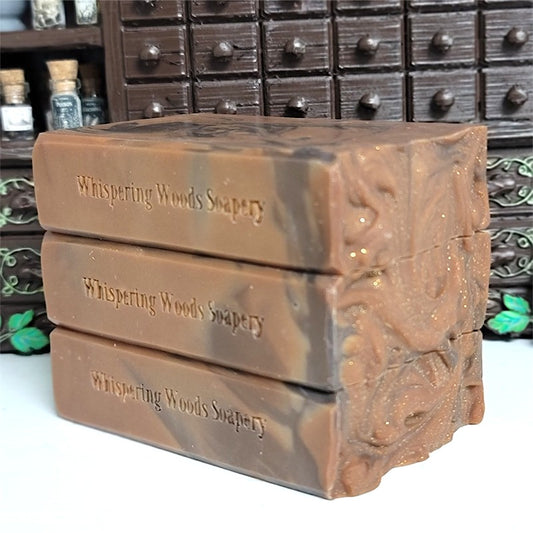 Three stacked rectangular bars of Whispering Woods Soapery soap in a burnt umber and brown color, with gold sparkle on top, set against a dark apothecary cabinet background.