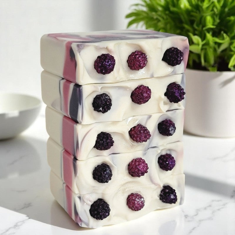 Five white soaps with purple and pink berries on top are stacked on a white marble surface with a plant in the background