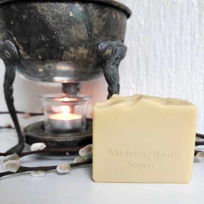 A rectangular bar of creamy off-white soap with 'Whispering Woods Soapery' stamped on it, set in front of a vintage liquid warmer with a lit tealight candle in the base.