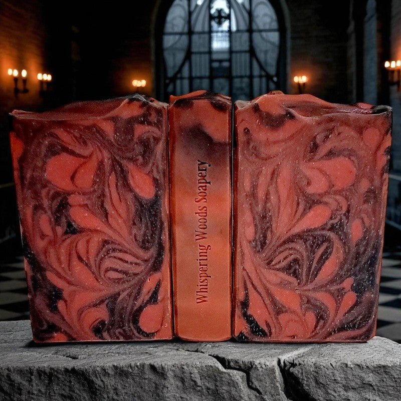 Three soap bars with red and black swirls and 'Whispering Woods Soapery' branding on a stone surface with lit candle sconces on the walls behind them