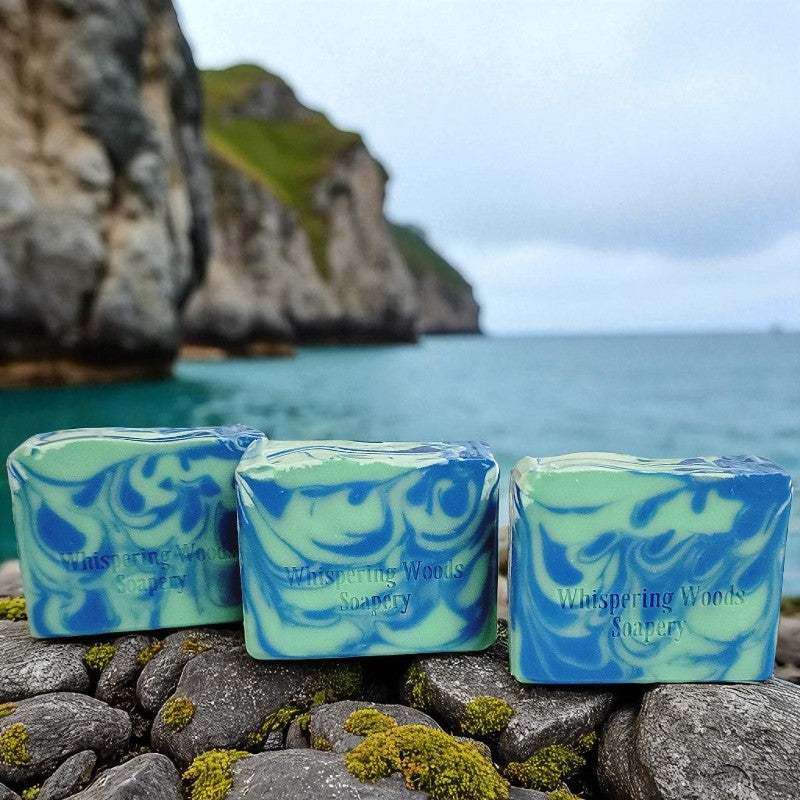 Three bars of 'Whispering Woods Soapery' soap in hues of blue and green sit on rocks with a scenic ocean background