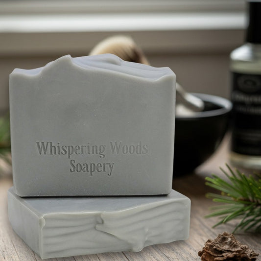 Silver bars of soap with 'Whispering Woods Soapery' branding on a wooden surface
