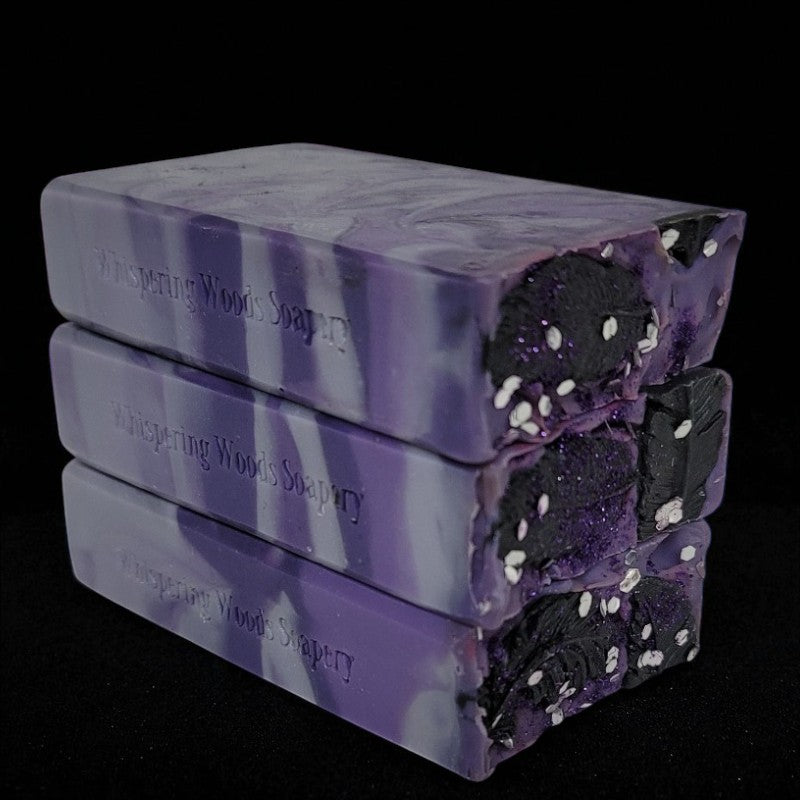 Stack of purple, silver and black soap bars with 'Whispering Woods Soapery' branding on a black background