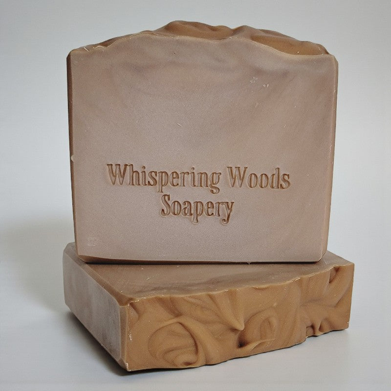 Two bars of light brown soap with 'Whispering Woods Soapery' branding on a light gray background