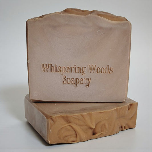 Two bars of light brown soap with 'Whispering Woods Soapery' branding on a light gray background