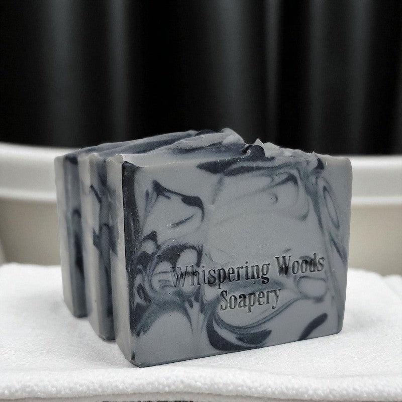 Three bars of gray and black soap with 'Whispering Woods Soapery' branding on a white surface with a black curtain background