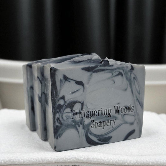 Three bars of gray and black soap with 'Whispering Woods Soapery' branding on a white surface with a black curtain background