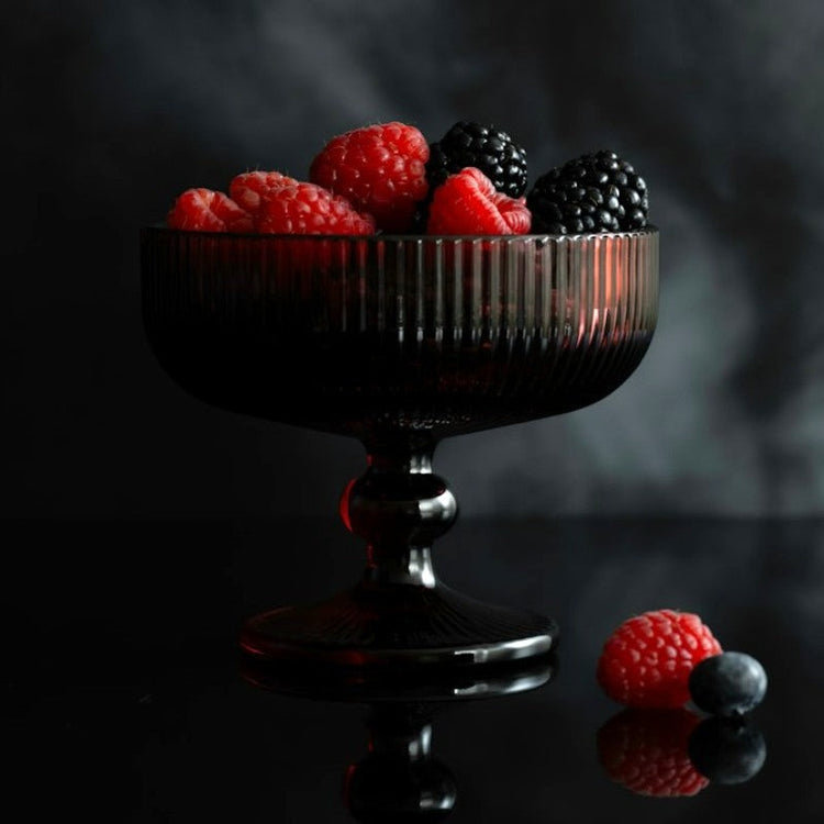 A dark chalice filled with blackberries and raspberries on a dark glossy table with a marbled gray background