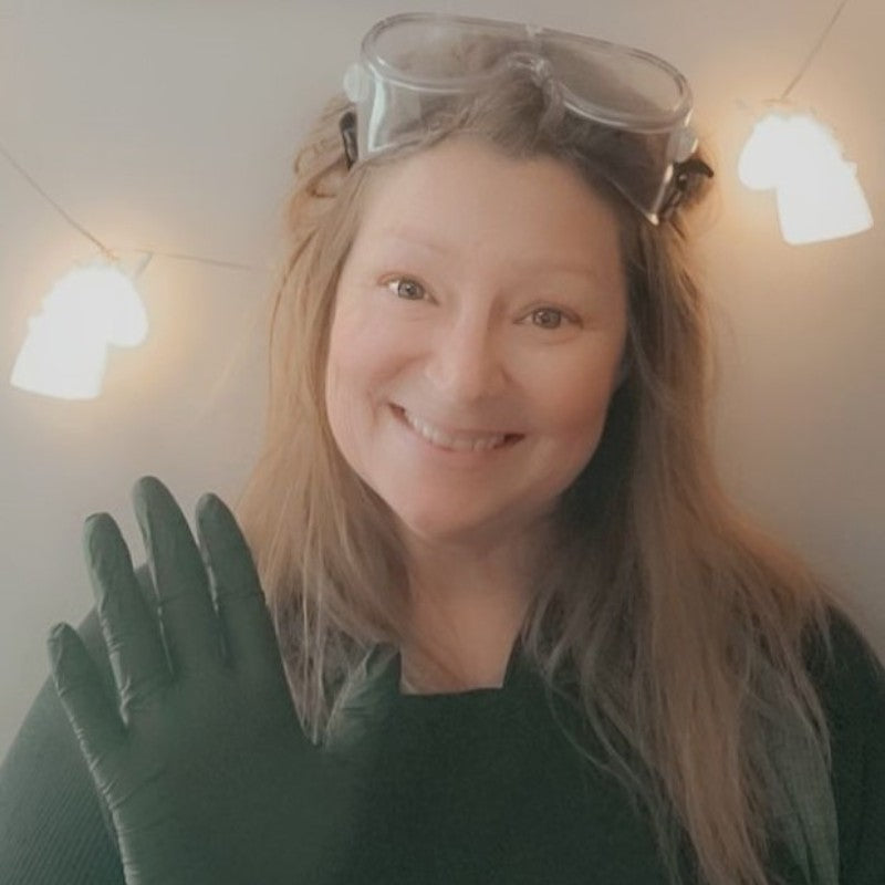 Misfit owner/soapmaker of Whispering Woods Soapery. She is waving with gloved hands. There are googles on her head, and small unicorn lights in the background.